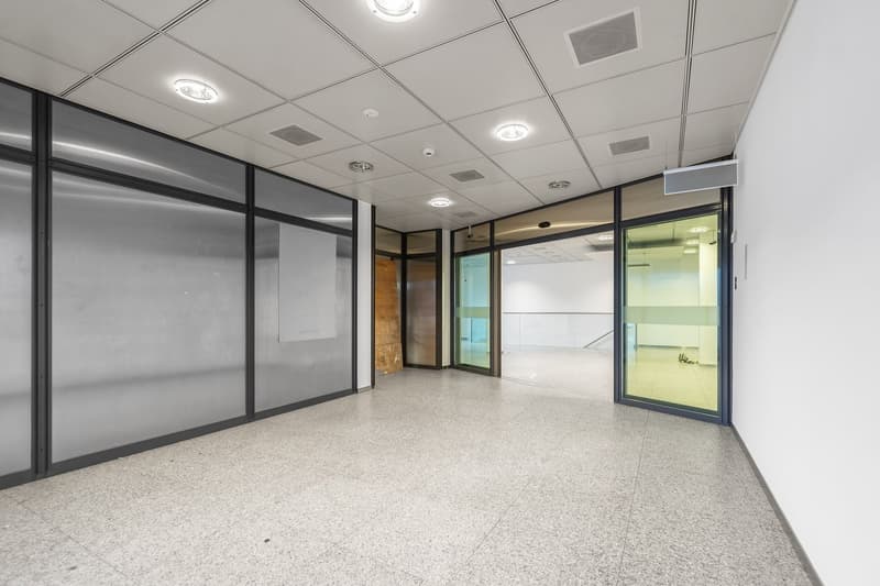 The image shows a large, open office space with a tiled floor and a suspended ceiling with recessed lighting. The space has glass walls and doors, creating a modern and professional atmosphere. There are no visible features from the provided list.