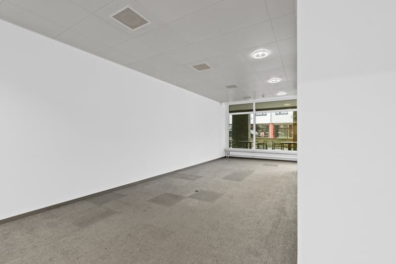 The image shows a large, open office space with a high ceiling, recessed lighting, and a tiled floor. The space appears to be empty, with no furniture or other objects visible. The windows at the far end of the room provide natural light and a view of the