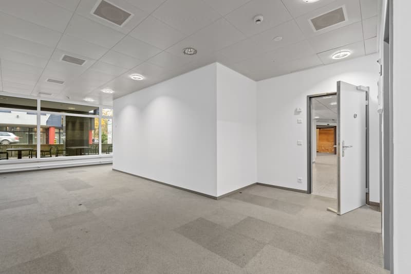 The image shows a large, open office space with white walls, a tiled floor, and recessed lighting in the ceiling. There is a glass door visible in the corner, leading to another room. The space appears to be empty and ready for use.