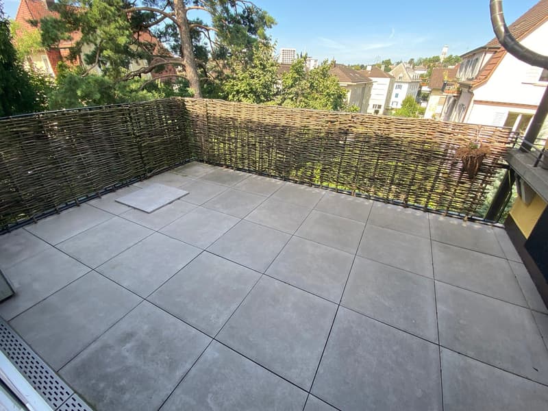 The image shows a balcony or terrace area with a woven wicker fence surrounding it. The floor is covered with large gray tiles. There are trees and buildings visible in the background, suggesting this is part of a residential or apartment building.