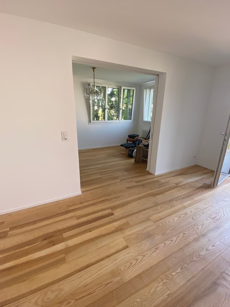 Spacious living room with hardwood floors, white walls, large windows, and a chandelier.