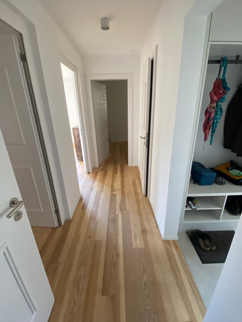 The image shows a hallway with white walls and a hardwood floor. The hallway leads to several rooms, with doors visible on the left and right sides. There is a closet or storage unit visible on the right side of the hallway.