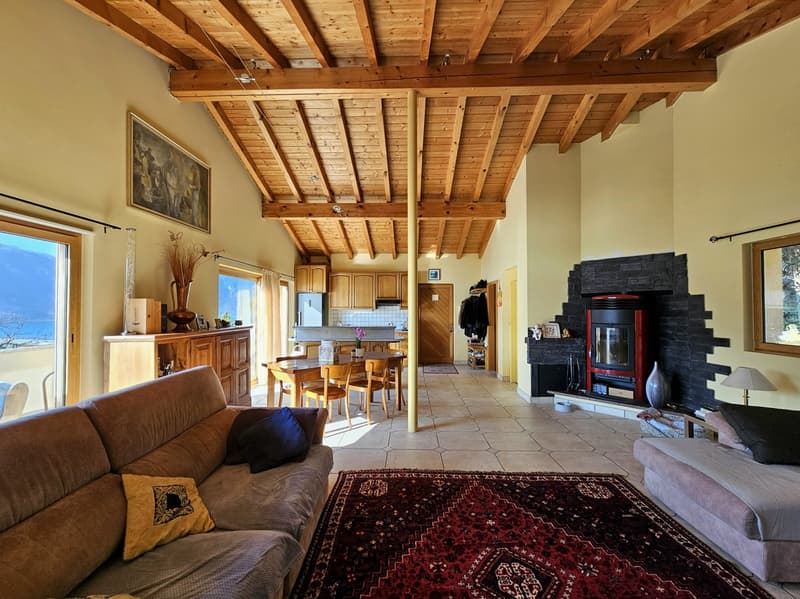 Open-concept living room and kitchen with exposed wooden beams on the ceiling, hardwood floors, a large brown sofa, a Persian-style rug, and a wood-burning fireplace with a black stone surround.