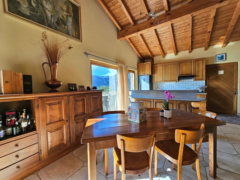Spacious open-plan kitchen and dining area with wooden beamed ceiling, wooden cabinets and furniture, large windows providing natural light, and a fireplace visible in the background.