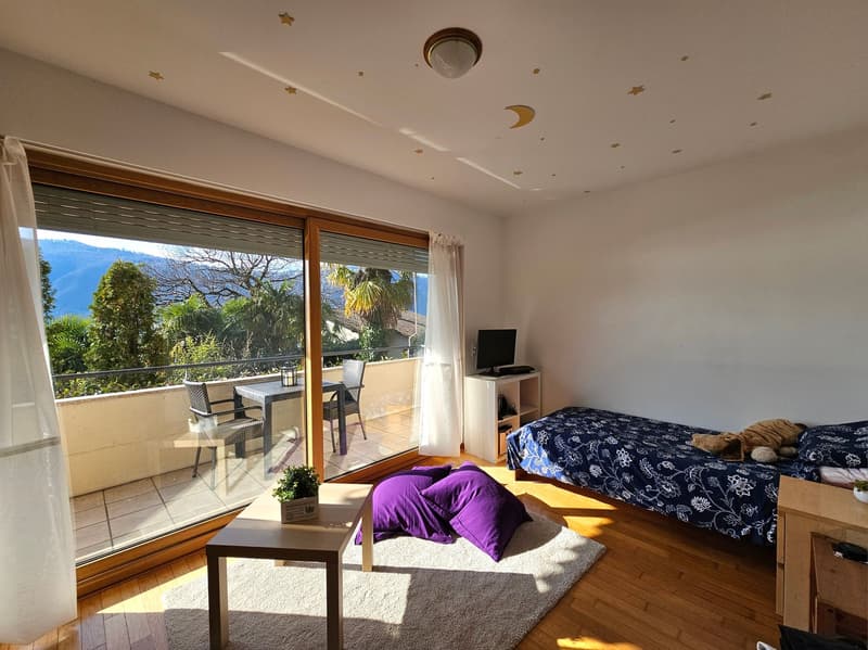 Spacious bedroom with large windows overlooking a balcony and scenic outdoor views. The room features a bed, a small table, and a TV stand. The balcony can be accessed through the sliding glass doors, providing access to an outdoor seating area.