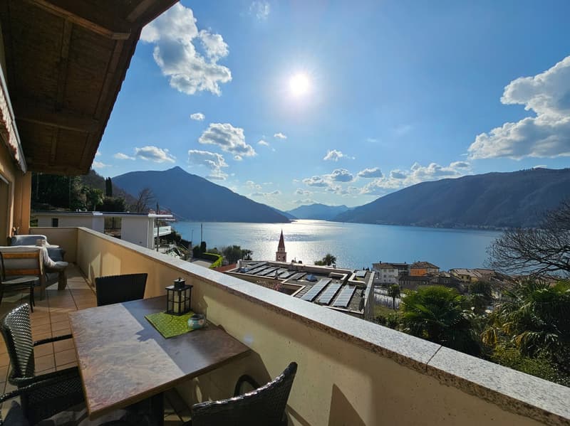 Balcony overlooking a scenic lake surrounded by mountains, with a sunny sky and clouds. The balcony has a table and chairs, and there is a small town visible in the distance.