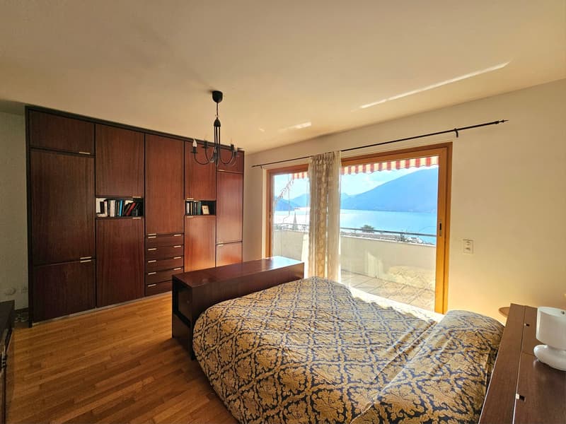 Spacious bedroom with wooden built-in wardrobes, a large bed with patterned bedding, and a sliding glass door leading to a balcony with a scenic view of the lake and mountains.