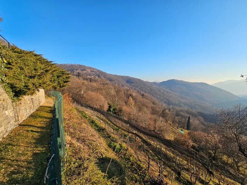 The image shows a scenic mountain landscape with rolling hills, forests, and a clear blue sky. There is a winding path or trail along the side of the hill, with a metal fence or railing running alongside it. The landscape appears to be in a rural or mount