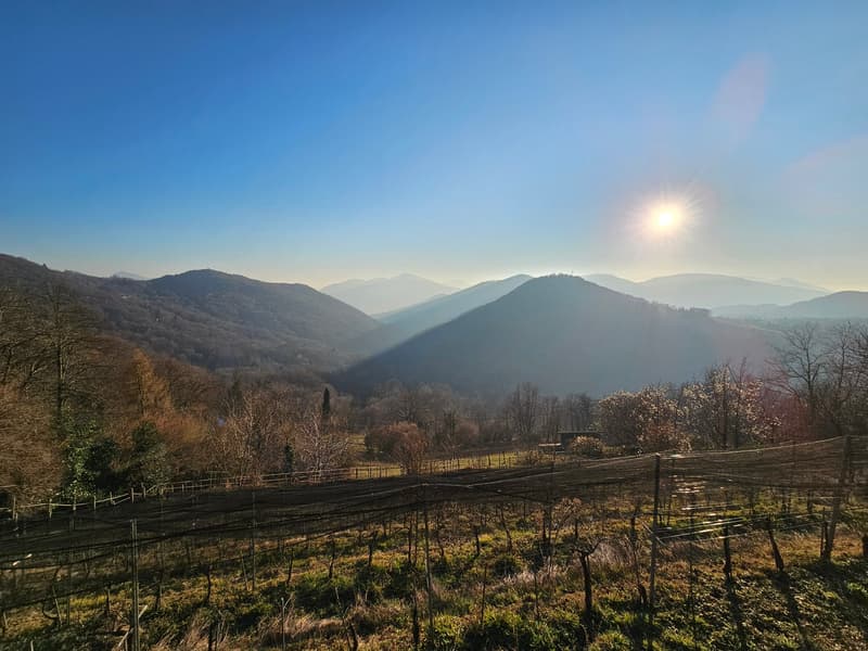 The image shows a scenic mountain landscape with rolling hills and valleys. The sky is a deep blue with some hazy clouds, and the sun is shining brightly. In the foreground, there is a fenced field or pasture, and in the distance, there are several mounta