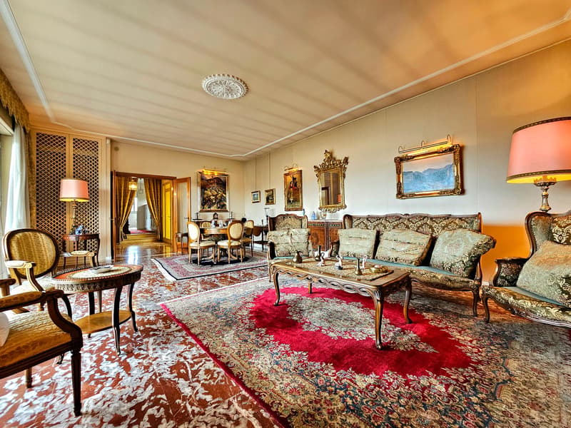 large living room, wooden floor, antique furniture, chandelier, paintings, mirrors, carpet, lamps, center table, armchairs, side tables