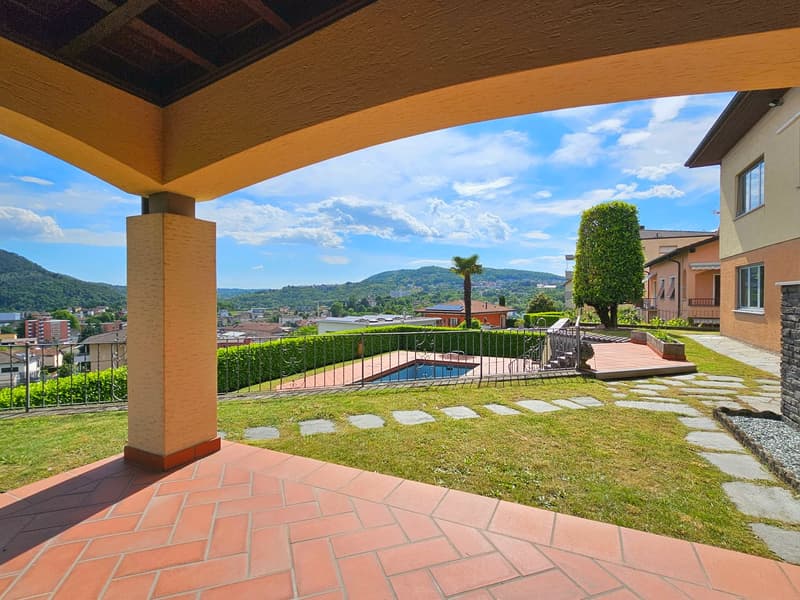 Patio with brick floor, covered by a wooden pergola, a swimming pool, and a garden. Mountains and a city landscape in the background.