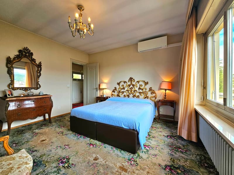 large double bed, blue linen, nightstands with lamps, mirror, chandelier, wooden cabinet with drawers, radiator, floral carpet, window with view