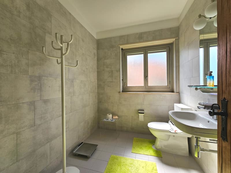 Small bathroom with tiled walls, white toilet, white sink, green rugs, window