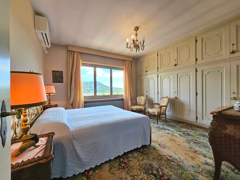 large bed, white coverlet, white curtains, ceiling chandelier, 2 armchairs, wooden cabinet, window with scenic view