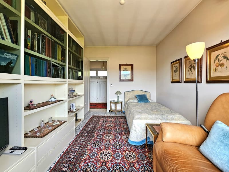 A room with a bed, shelves with books, carpet, a chair, a side table, and a lamp.