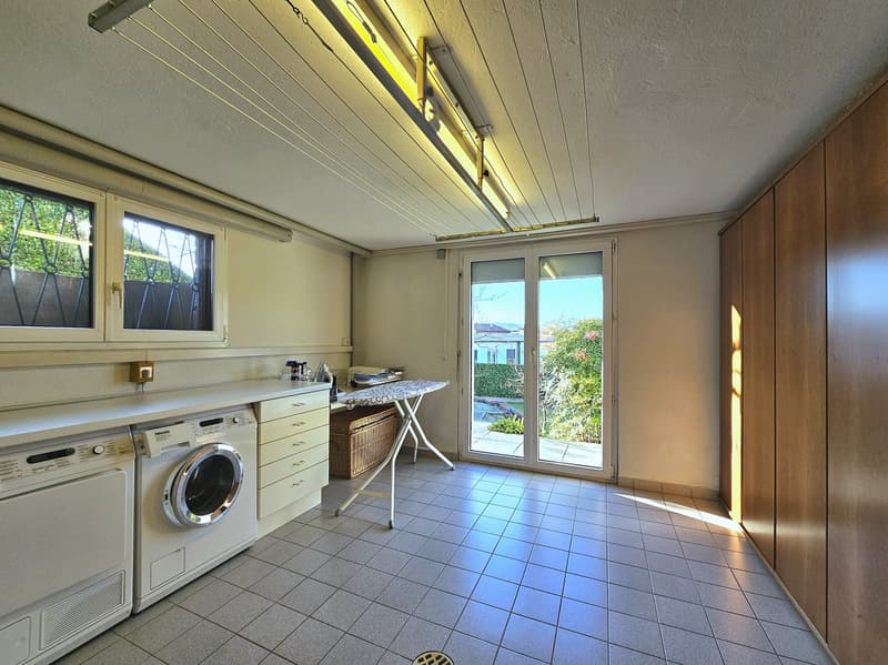 A laundry room with tile flooring, a washing machine, dryer, ironing board, a basket, and a cabinet