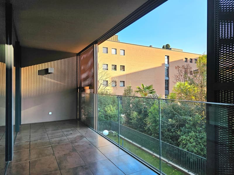 Balcony with glass railings, tiled floor, open view to the outside, plants and trees visible