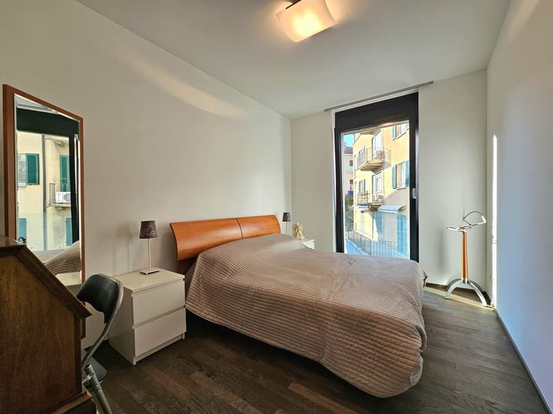 Bedroom with bed, nightstands, lamps, mirror, and a chair. Large window with a view of the outside.