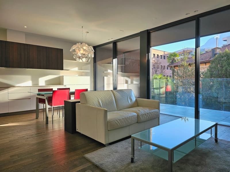 modern living room with white leather couch, glass coffee table, glass doors leading to balcony, wooden floor, dining table with red chairs