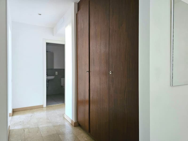 Empty room with white walls, wooden floor, large wooden closet, mirror, and a bathroom with a toilet and sink