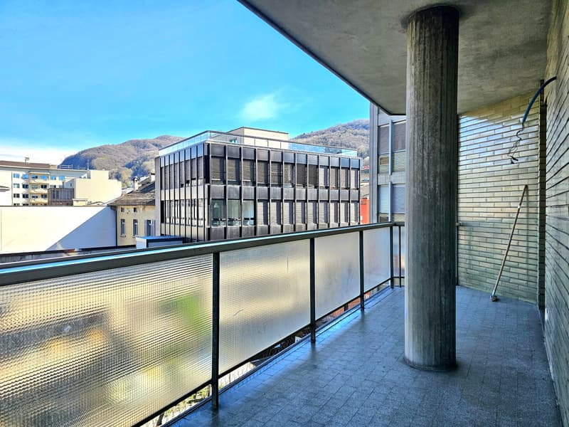 Balcony with glass railing, tiled floor, central column, distant view of mountains and buildings