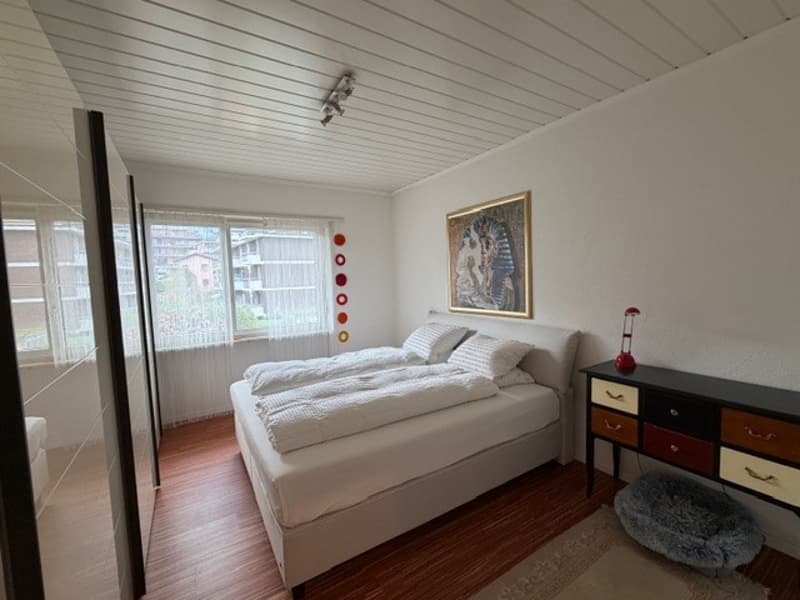 A bedroom with a bed, nightstand, and rug. The bed has white sheets and pillows. The nightstand has a lamp and a drawer. The room has a white wall and a wooden floor. There is a painting on the wall and a mirror on the left side. The window has white curt