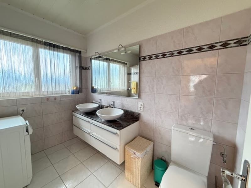 Bathroom with two sinks, a toilet, a washing machine, and a cabinet. The room has tiled walls and floors, and a mirror above the sinks.