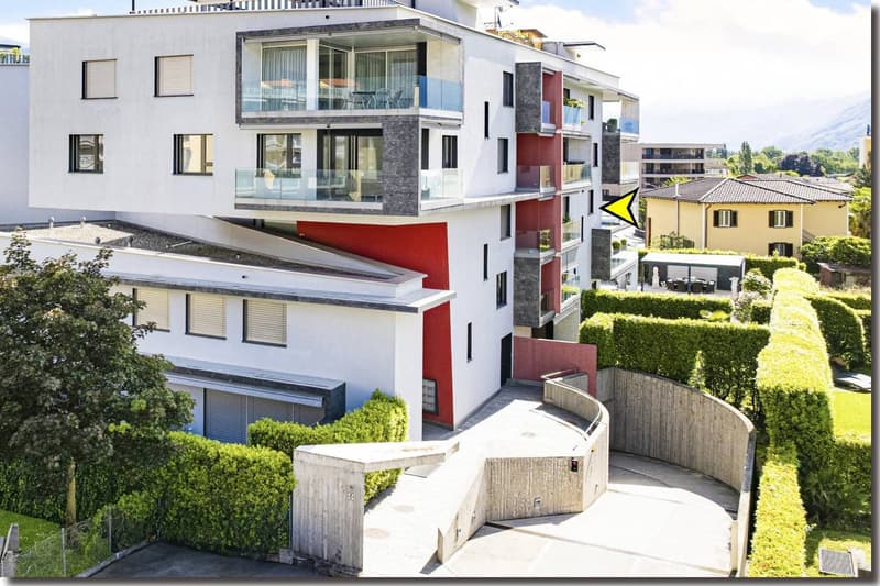 Modern apartment building with multiple floors, white and red facade, glass balconies, fenced entrance, hedges, and a small garden