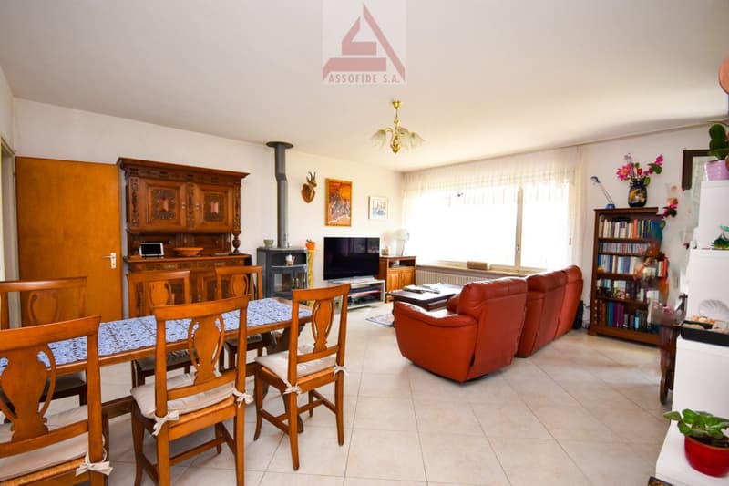 Spacious living room with wooden furniture, including a dining table and chairs, a large cabinet, and a red leather sofa. There is a fireplace visible in the room, and a TV stand with a TV. The room also has bookshelves and decorative items.