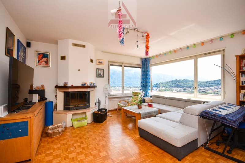 Spacious living room with large windows offering a scenic view, hardwood flooring, fireplace, sofa set, coffee table, and various decorative elements like colorful garlands and framed artwork on the walls.