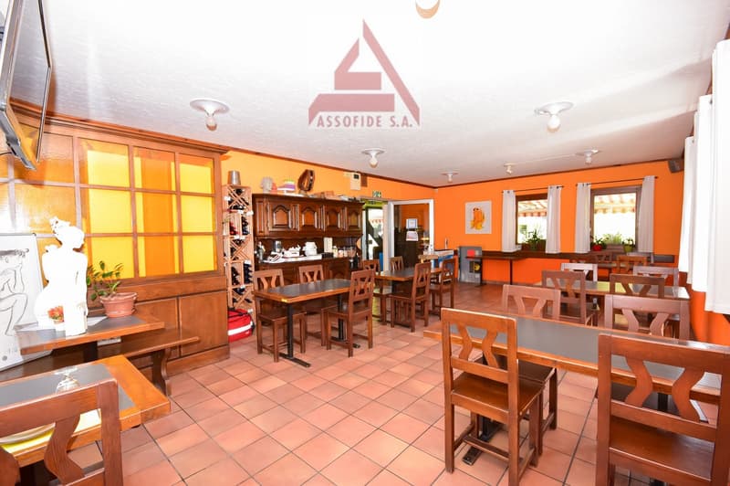 The image shows the interior of a restaurant or cafe. The space has an orange color scheme with wooden furniture, including tables and chairs. There is a counter area with shelves and decor items. The ceiling has recessed lighting fixtures. The overall at
