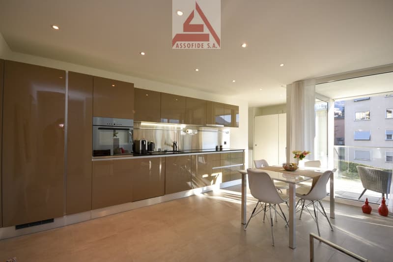 Spacious open-plan kitchen with modern, high-gloss brown cabinets, stainless steel appliances including a dishwasher, and a dining area with a table and chairs next to large windows providing natural light.