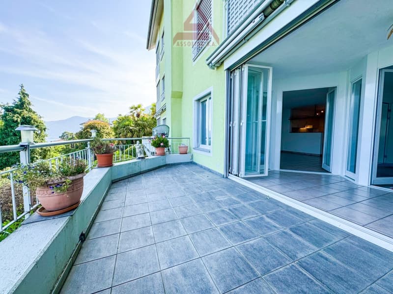 The image shows a balcony or terrace of an apartment. The balcony has a tiled floor and is surrounded by a railing. There are potted plants and flowers on the balcony. The apartment building has large windows and a green exterior.