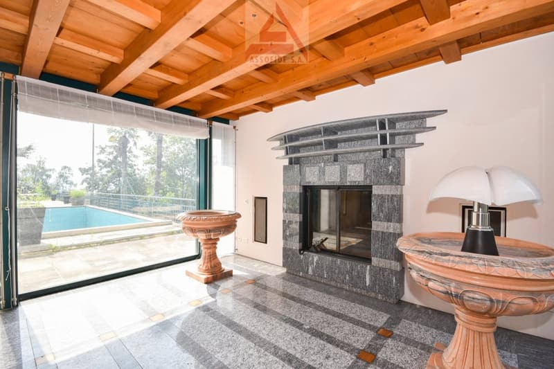 Spacious living room with wooden beamed ceiling, large windows overlooking a swimming pool, a stone fireplace, and decorative ceramic vases.