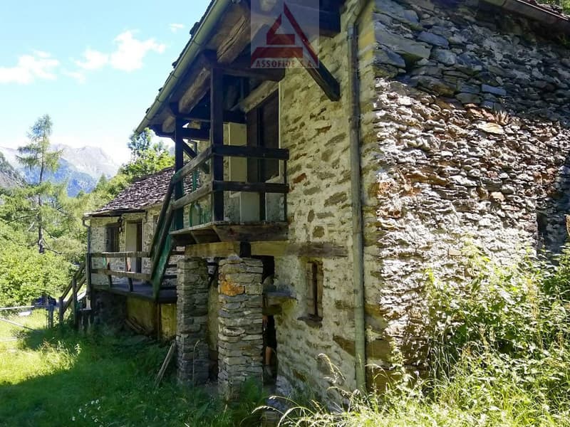 A rustic stone building with a wooden balcony or terrace, surrounded by lush greenery and mountains in the background.