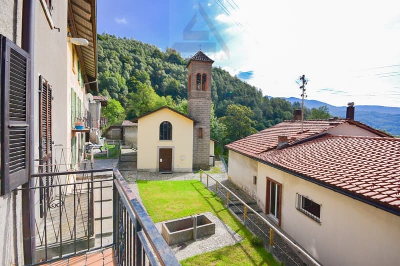 The image shows a picturesque village nestled in a lush, green mountainous landscape. The buildings have traditional red tile roofs and there is a small church with a bell tower. The scene includes a balcony or terrace overlooking the village and the surr