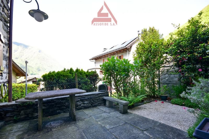 Outdoor seating area with a stone bench, a table, a small garden with flowers, plants, and a stone wall. There is a house in the background.