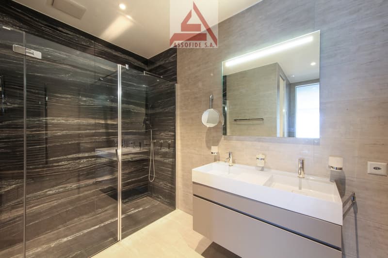A modern and sleek bathroom with a glass walk-in shower, marble-style tiles on the walls, a white vanity with a double sink, and built-in wall-mounted mirror. The walls are finished with light-colored tiles, and the floor is wooden. The room is illuminate