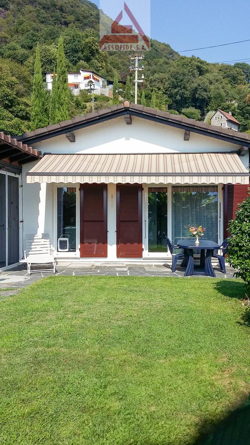 detached house, brown tiled roof, white exterior, awning, glass doors, table and chairs, grassy lawn, distant house, wooded mountain
