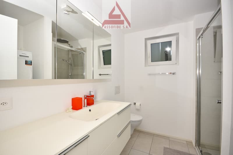 Modern bathroom with white cabinets, a white sink, a glass shower, and a toilet. The room includes a window and a mirror.