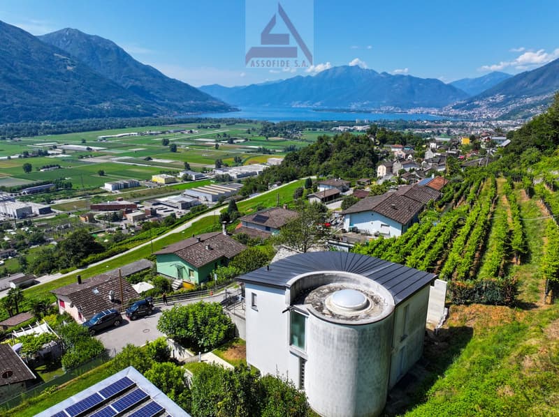 a modern house with a cylindrical tower in the center, surrounded by houses, vineyards, a lake, and mountains
