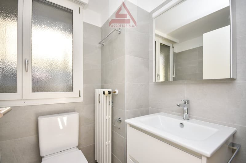 White toilet, white sink, white cabinet, grey tiles, window, white radiator