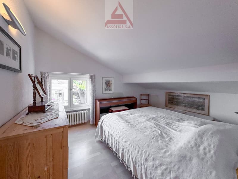 light colored room, large bed, wooden dresser, window with curtains, white sheets, hardwood floor, radiator, picture frames