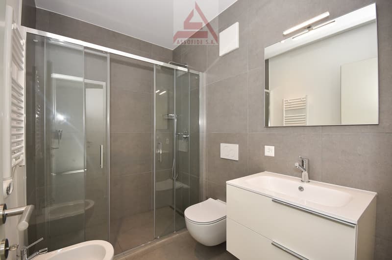 Bathroom with glass shower, white ceramic sink, white cabinets, white toilet, mirror, and wall-mounted lights