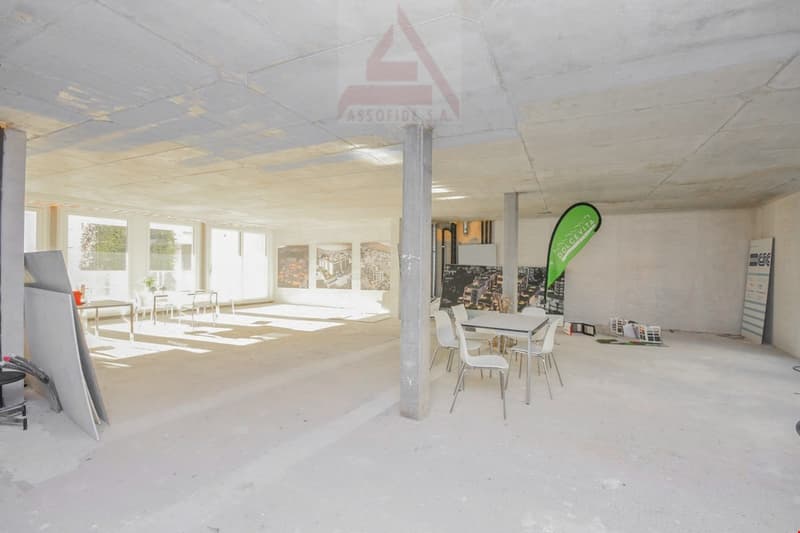 Unfinished basement space with white walls, grey concrete floor, large windows, central support pillar, some empty tables and chairs, and architectural promotional banners
