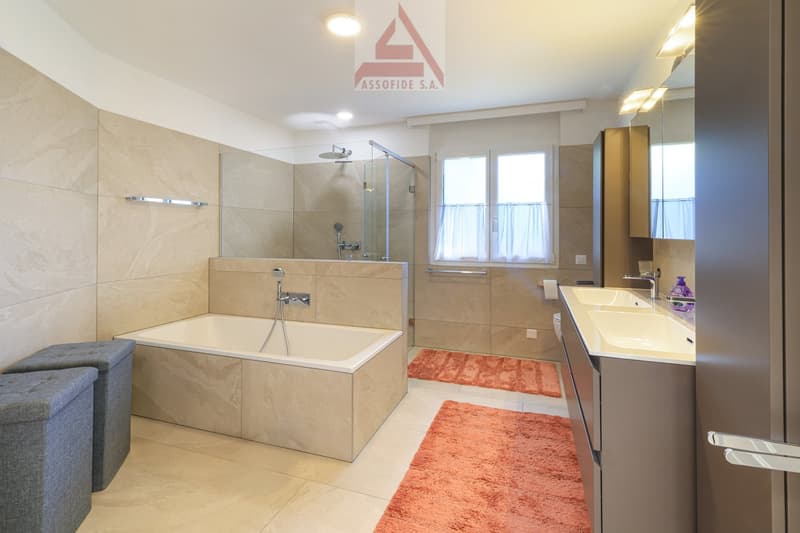 Bathroom with bathtub, shower, double sink, and mirror. Tiled walls, two rugs, two window with curtains.