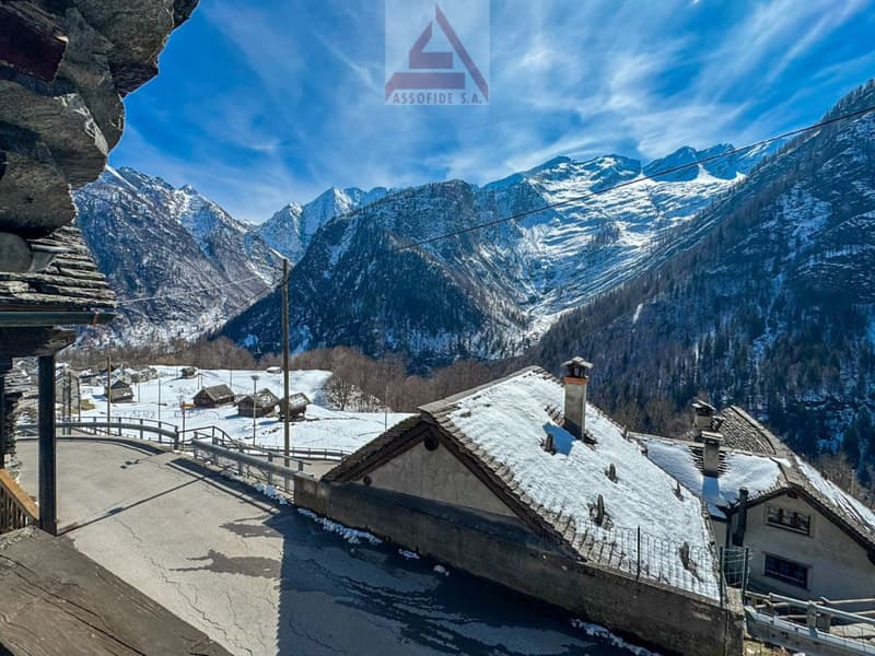 Mountain house with snow-covered roof, stone walls, chimneys, surrounded by snowy mountains and a clear blue sky