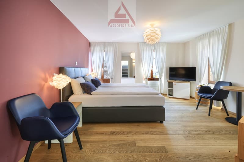 Bedroom with double bed, pink wall, wooden floor, white curtains, lamps, TV, wooden table, blue chairs, wooden barrel