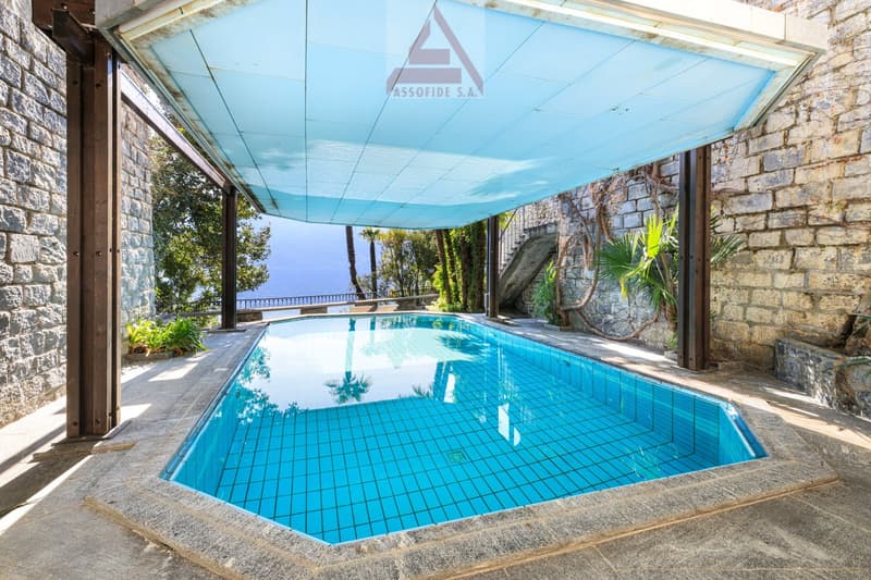 Outdoor swimming pool with blue tiles, covered by a blue canopy, surrounded by stone walls, with a view of the sea and greenery.