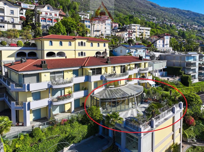 Aerial view of a multi-story building with red roofs, yellow walls, multiple balconies, and a highlighted glass terrace with a view of the mountains.